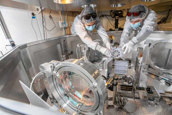 Photo of two people in clean room equipment leaning over a complex array of lenses and electronic equipment