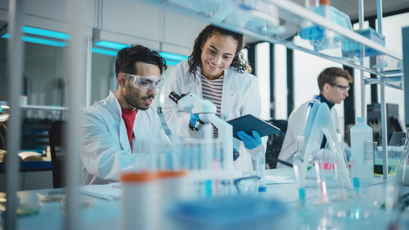Stock photo of a Hispanic man at a microscope and a Black woman next to him, both wearing lab coats