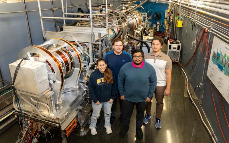 Photo of  Isabelle Dib, Dominik Dziura, Stuart Castillo and Maksymilian Dziura standing next to the Neutron Spin Echo spectrometer