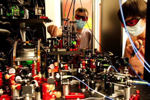 Photo of two people in lab goggles and face masks working with a lab bench full of optical and quantum measurement equipment