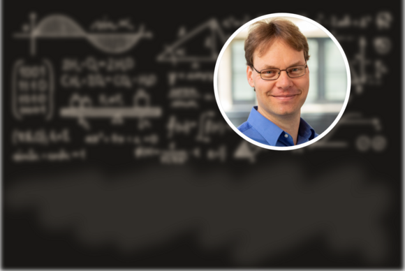 Photo of Rouven Essig (a white man in a blue shirt) against a background of a blackboard with blurred equations