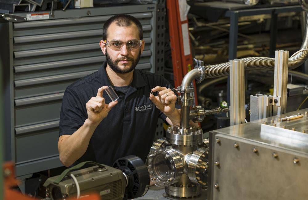 Photo of an engineer (a white man) holding up a small plastic canister, sitting behind a tool at the Fuel Pellet Fueling Laboratory