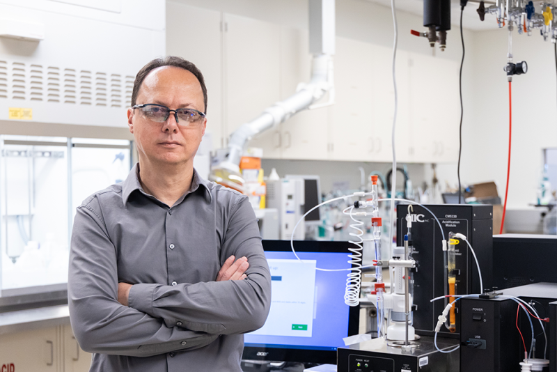 Photo of Radu Custelcean (a white man in a button-down shirt) standing in front of a computer and scientific equipment