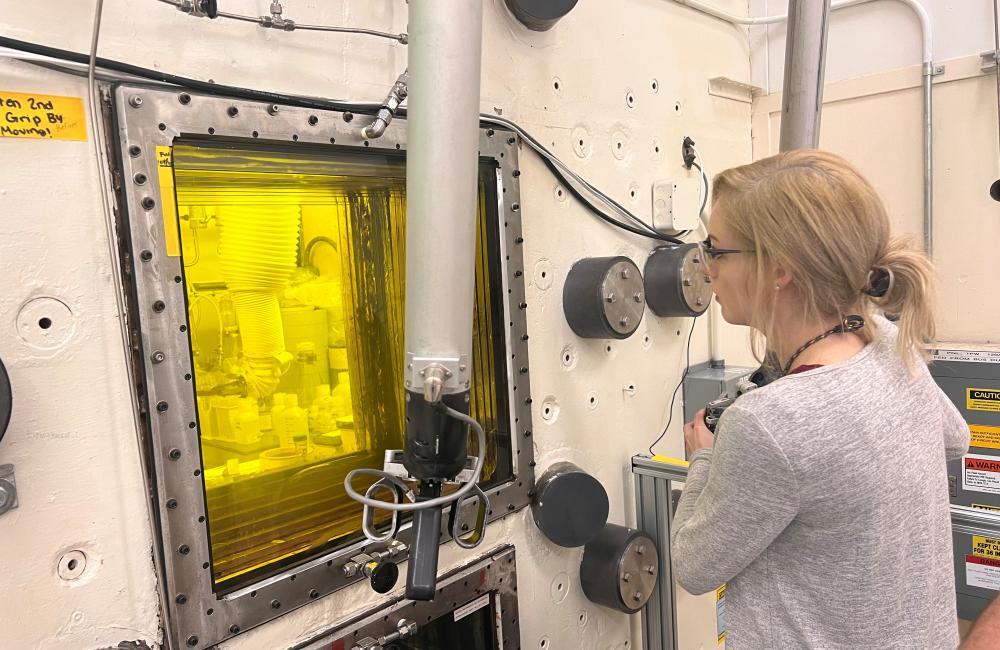 Raina Setzer (a white woman in a gray shirt) operating equipment within a hot cell that has a yellow color inside