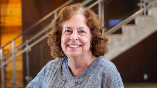 Beverly Marzec (a white woman in a grey sweater) sitting in a lobby with a staircase behind her