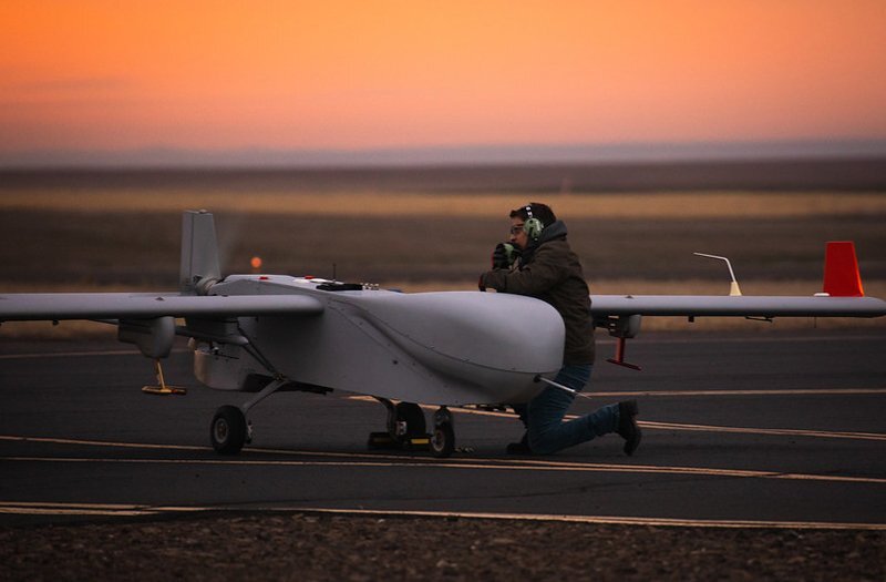A pilot kneels next to a small unmanned aerial system, a small, white plane-like device on a runway with a sunrise in the background