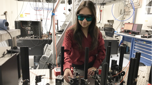 Chloe Washabaugh (a white woman in lab goggles) working with a quantum tech set-up on a lab bench with more equipment behind her