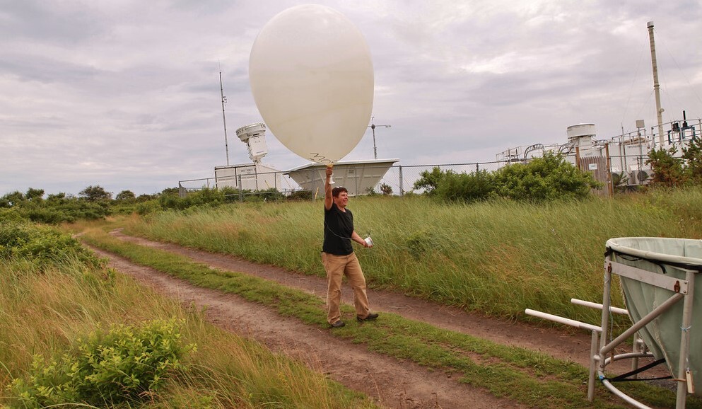 A scientist (a white man) holding a large weather balloon in a field with a dirt track through it and atmospheric measurement equipment behind him