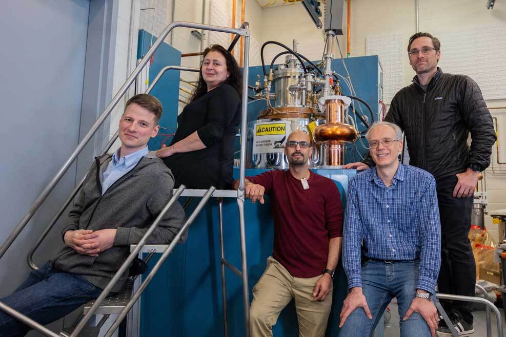 Group of five scientists (four men, one woman) sitting around particle accelerator equipment