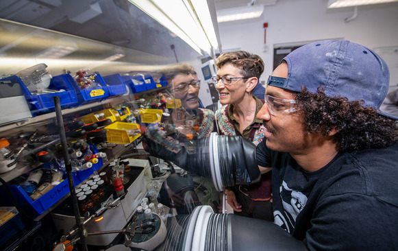Polly Arnold (a white woman in an olive shirt) standing next to Matt Hernandez (a Hispanic man in a t-shirt), who is using a glove box in a lab.