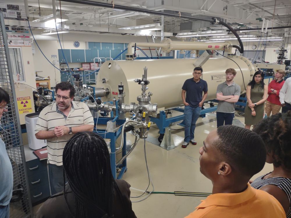 A group of students of various races and genders watching a Latino man speak in a laboratory with a large cylindrical piece of equipment
