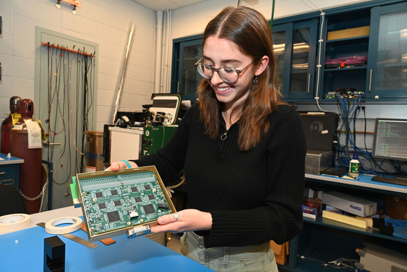 Nora Lowe holding up a board of microelectronics, standing in a laboratory with a shelf of electronics behind her.