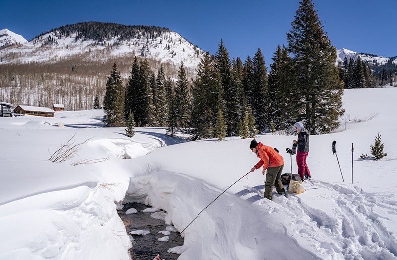 Researchers in winter clothes taking a sample from a stream surrounded by snowy land, with mountains in the background