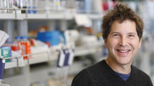 David Baker (a white man in a black sweater) standing in a biochemistry lab with equipment behind him