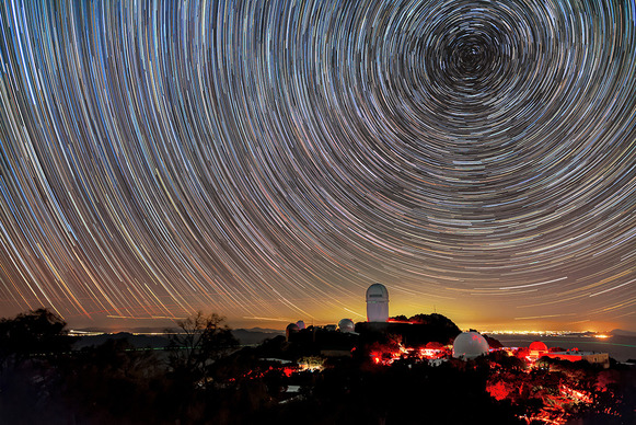 A observatory on a hill with glowing lights below and a swirl of long-exposure stars above