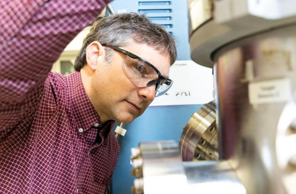 Gabriel Veith (a man in lab goggles and a checked shirt) looking at a metallic piece of scientific equipment