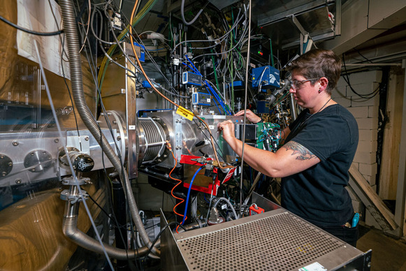 Jacklyn Gates (a woman in a black t-shirt) working on a metallic piece of machinery with wires coming out of it