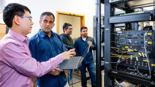 Four researchers (all men) in a laboratory with a rack of electronics equipment. Two of them are looking at a laptop.