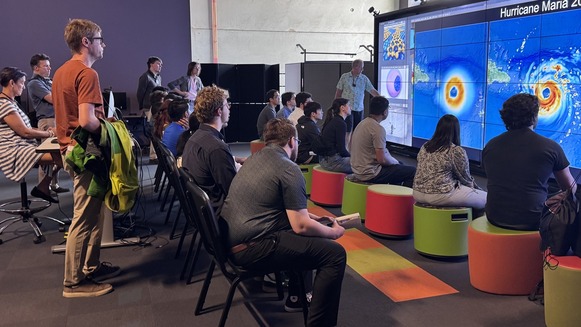 Group of people sitting on multi-colored seats and stools looking at a large screen showing images of Hurricane Maria