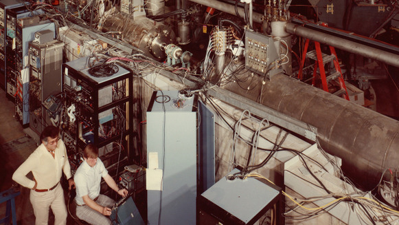 Historical photo of Paul Markovich and Gene Clifft testing the booster section of ATLAS, made up of metal cylinders with wires and electronics.
