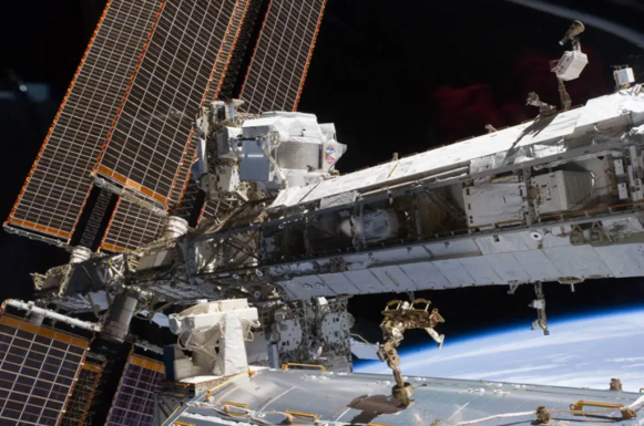The tube of the International Space Station runs from left to right. The AMS-02 is a white box-shaped machine in front of the solar panels.