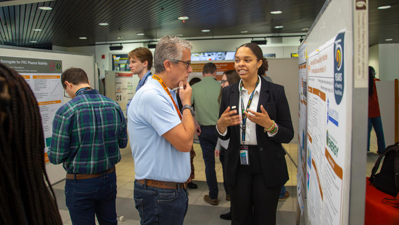 A woman in a suit standing next to a scientific poster, explaining what is on the poster to a man looking at the poster