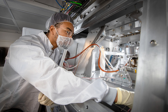 A man in a clean room suit working with manufacturing equipment under a lab hood