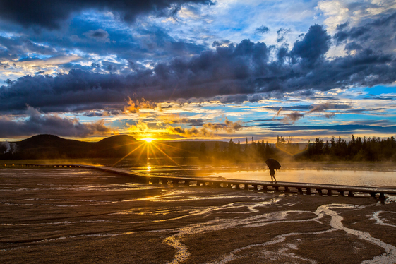A lone visitor walks across the path over the thermal springs as the sun rises over the mountains in the background. 
