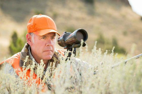 Secretary Zinke wearing hunting gear and sitting in high grass with a large scope.
