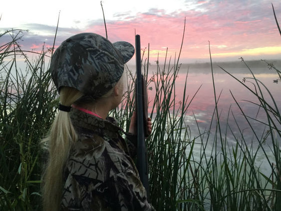 A young woman in camouflage hunting gear holds a shotgun and stands in tall grass looking at a pink sunrise.