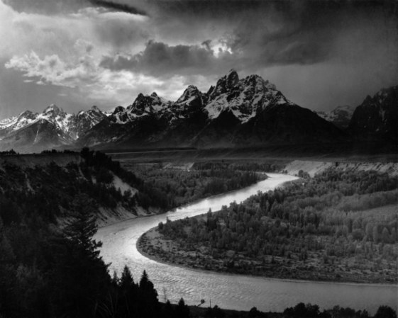 A black and white photo of a river curving around some trees with a range of tall jagged mountains on the horizon.