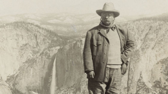 Historic black and white photo of Theodore Roosevelt in a hat, coat and glasses with a view of mountains and a waterfall behind him.