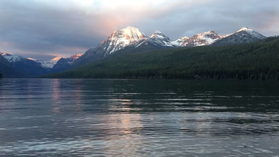 The sun sets over a rippling lake with tall snow-capped mountains in the background.