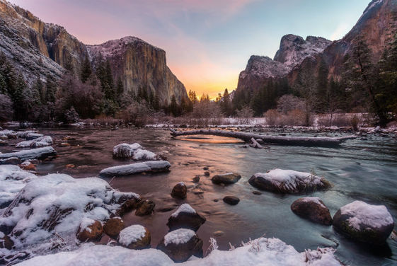A narrow creek flows over rocks in a wide valley dusted with snow and bordered by high rocky cliffs at sunrise.