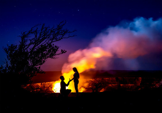 A man kneels on one knee and proposes to a woman next to a flaming volcanic crater under a dark, starry sky.