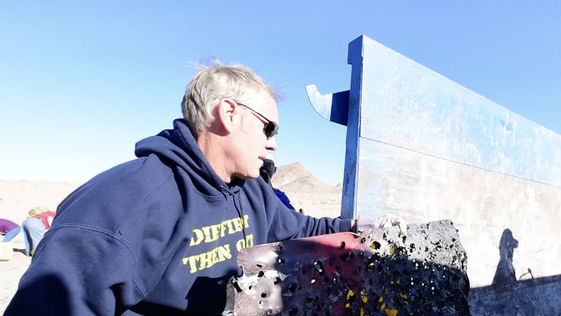 Secretary Zinke, a white man with shirt, gray hair wearing a sweatshirt, lifts a large piece of metal trash into the back of a truck.