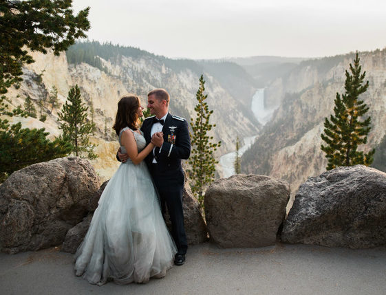 Photo of a woman in a wedding dress and a man in a military uniform smiling at each other as they stand on a path over looking a waterfall.