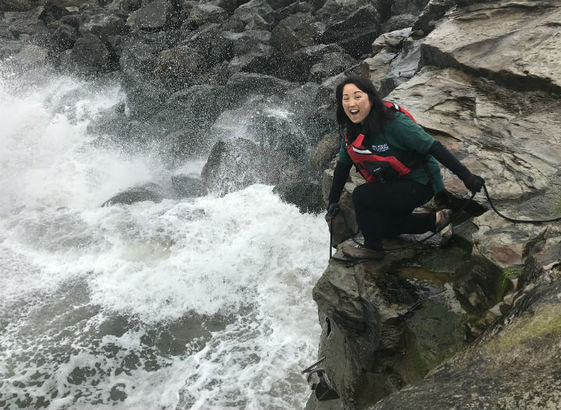 A smiling Asian American woman in a USGS shirt and safety gear kneels on a rock and lowers a metal scientific instrument into a river below her.