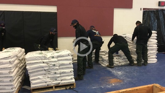 Five people in black clothes and masks stack large bags on a loading dock.