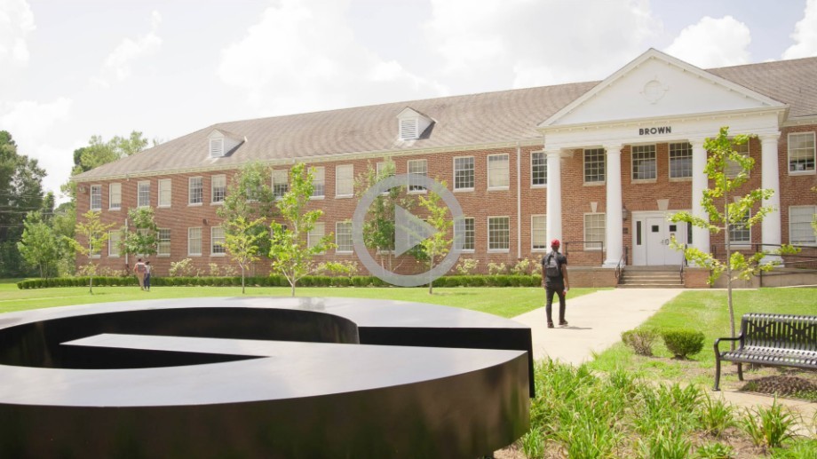 A young black man wearing a backpack walks in to a large brick building on a college campus.