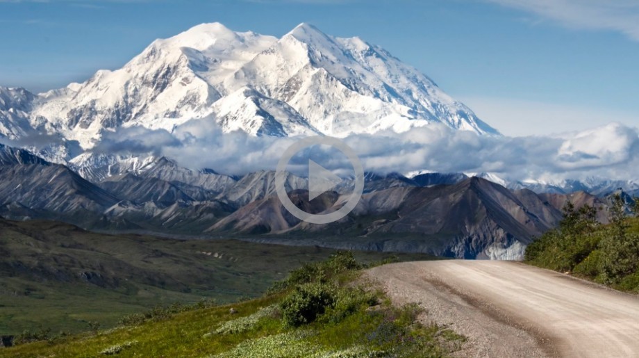 A dirt road leads over a hill towards a giant snow covered mountain.