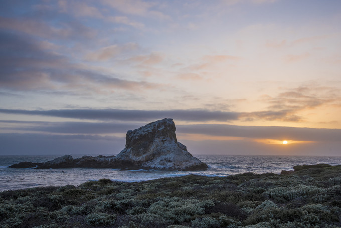 Rock protruding from the Pacific ocean near the shore with the sun setting in the background.