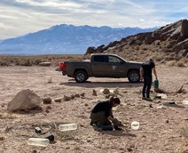 A truck parked with two people working on the ground with mountains in the background.