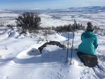 A person sitting on a rock in the snow facing a mountain range with ski poles in the snow next to her.
