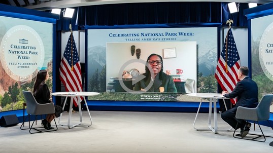 Secretary Haaland and the Second Gentleman address a park ranger on a large television screen