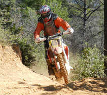 An off-highway dirt bike rider in the BLM Chappie-Shasta OHV Recreation Area on a dirt incline with dense brush and pine trees in the background.   