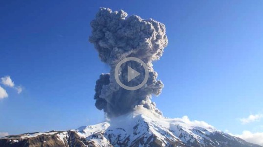 A snow covered volcano erupts a large plume of ash and smoke