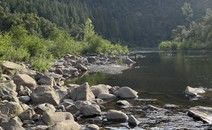 Expanse of American River lined with large river rocks, and pine covered hills. 