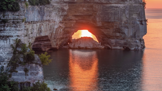 A bright orange reflection of the sun shines through a larch sandstone arch, reaching out into the water