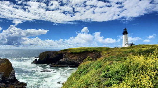 A lighthouse sits along the rocky coast of Oregon. Puffy white clouds are in a blue sky above